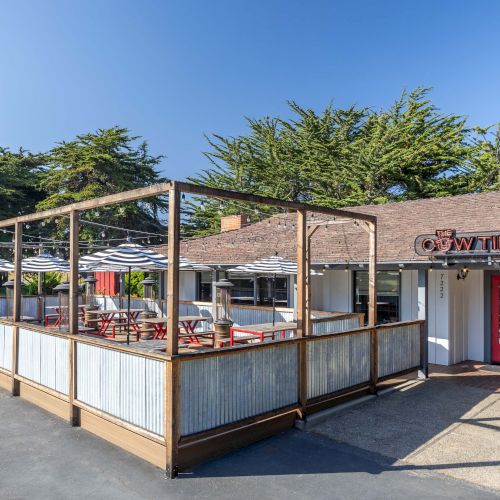 A small outdoor dining area with a covered wooden railing, adjacent to a brick building with red doors and a sign, sunny day.