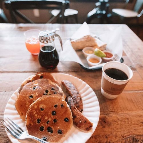 A cozy breakfast scene: pancakes with chocolate chips, sausage, a small pitcher of syrup, orange juice, coffee, and a pastry on a wooden table.