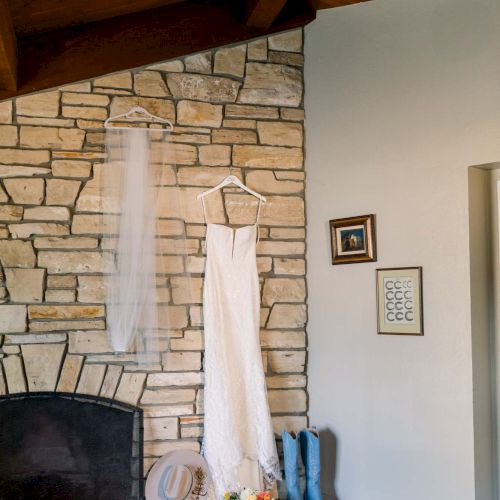 A bridal gown hangs on a wooden beam near a stone fireplace, with a veil and bouquet on the hearth, and blue shoes beside it, in a cozy room.