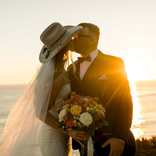 A bride and groom share a kiss at sunset on a cliff, the bride holding a bouquet, the veil flowing, romantic wedding moment.