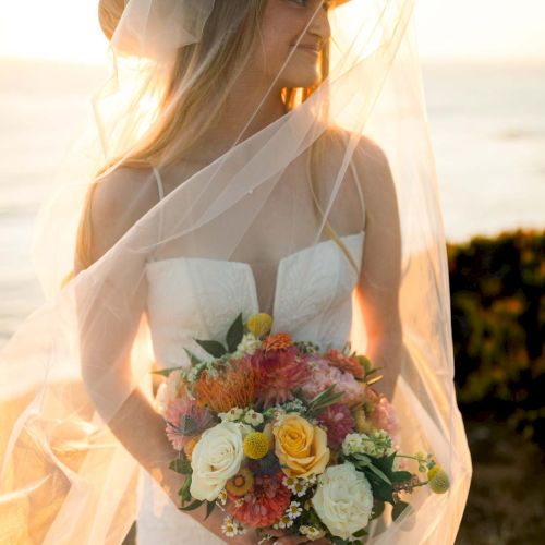 A bride in a white dress with a veil and hat stands in sunset, holding a colorful bouquet of roses and blooms by the shore.