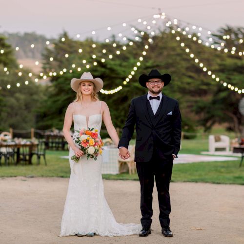 A couple in wedding attire walks hand-in-hand on a sandy outdoor venue, string lights overhead, bouquets and chairs set up in the background.