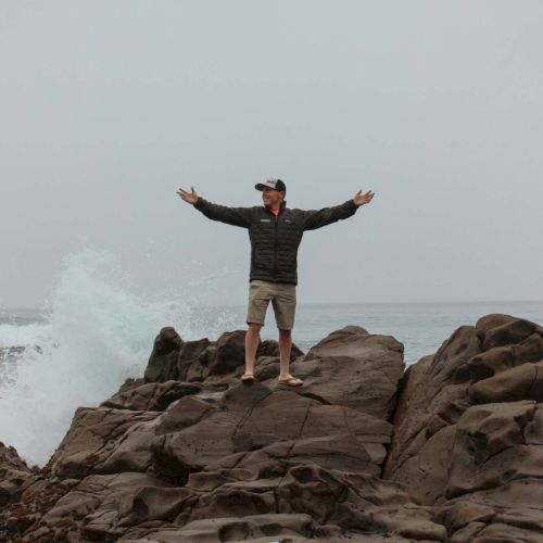 A person stands on jagged rocks by the sea with arms wide open, waves crashing behind, overcast sky, posing triumphantly.