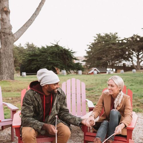 Two people sit in pink chairs by a fire pit, holding hands, chatting warmly on a grassy park with trees in the background.