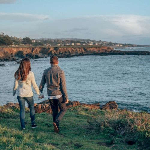 A couple walks hand in hand along a rocky shoreline with grass in the foreground and waves crashing against the coast at sunset.