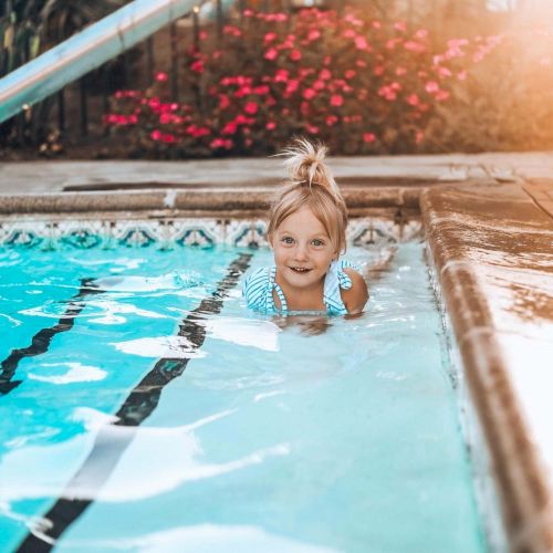 A happy young girl swims in a pool, smiling at the edge by the lane lines, with sunlight warm on the water and surrounding tiles.