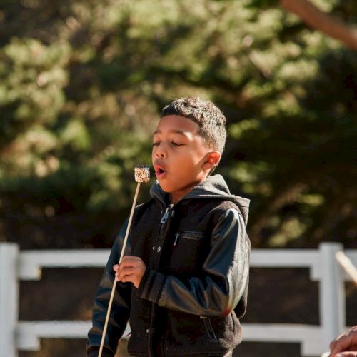 A young boy is speaking into a mic outdoors, likely at a school or event, wearing a black jacket and jeans, with trees in the background.