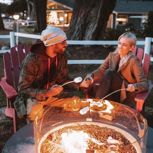 Two people sit around a fiery fire pit, chatting warmly in a cozy outdoor setting with chairs and trees at dusk.