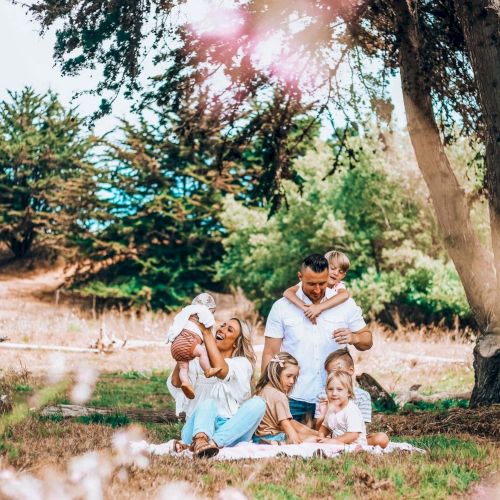 A family of six sits on the grass under trees, enjoying a sunny picnic in a park with blossoms and warm, joyful vibes.