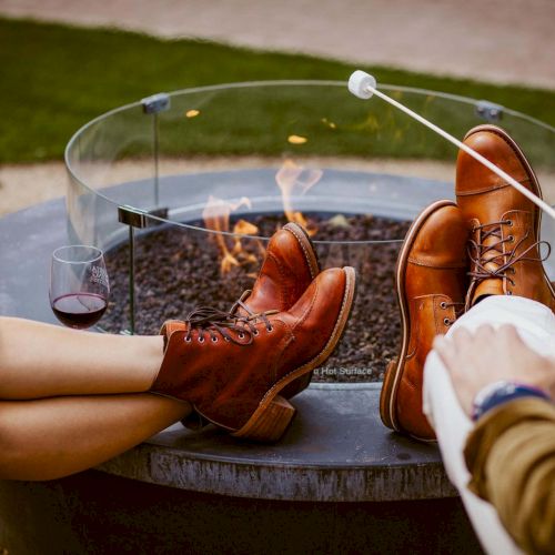 Two people with brown boots sit by a fire pit, roasting marshmallows on sticks and sharing a glass of wine nearby.