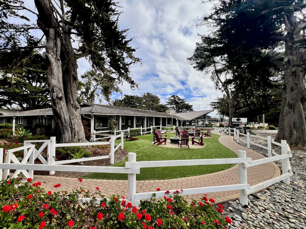 A well-kept outdoor courtyard with white fences, wooden benches, and flowers, set among tall trees near a low building, under a partly cloudy sky.