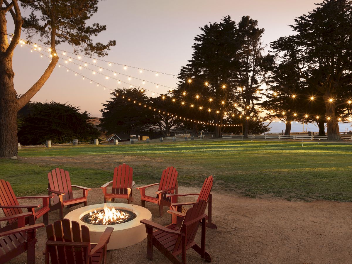 A cozy outdoor gathering with a circular fire pit surrounded by red Adirondack chairs, string lights overhead, trees, and a grassy park at dusk.